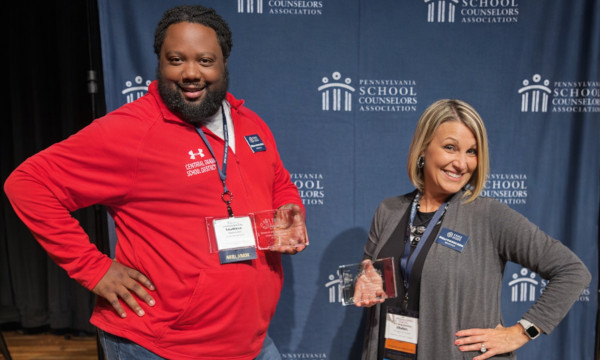 Two school counselors stand on stage holding glass awards in front of a blue Pennsylvania School Counselors Association