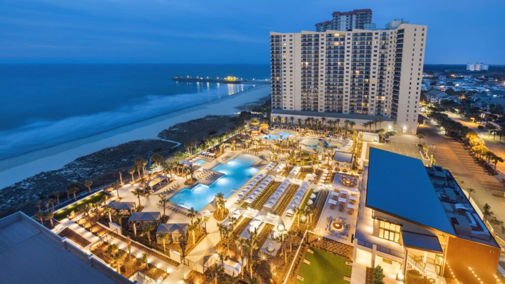 An overhead picture of the embassy suites on the beach at dusk, with a beach and pier in the background, and a large outside gathering area that includes a pool in the foreground.