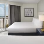 Hotel bedroom with two beds with a nightstand in between, and a glass door with a patio overlooking a beach.