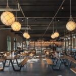 Modern restaurant interior with long wooden communal tables, benches, hanging globe pendant lights, dark ceiling beams, and large garage-style windows letting in natural light.