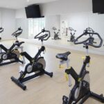 Large open fitness room with large windows on one wall and large mirrors and TV's on another wall, and several stationary bike machines.
