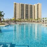 A large pool in the foreground surrounded by chairs on one side and palm trees on the other, with an Embassy Suites hotel in the background.