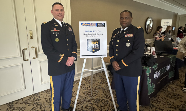 Two uniformed members of the North Carolina Army National Guard stand indoors beside a sign on an easel that reads “Thanks to our Silver Level and Opening Keynote Sponsor North Carolina Army National Guard.”