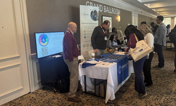 Attendees gather around a RethinkEd exhibition booth outside a room labeled “Grand Ballroom” at a conference. The booth features a blue tablecloth with the RethinkEd logo, informational materials, a laptop, and a banner promoting the “RethinkEd Wellness Suite.”