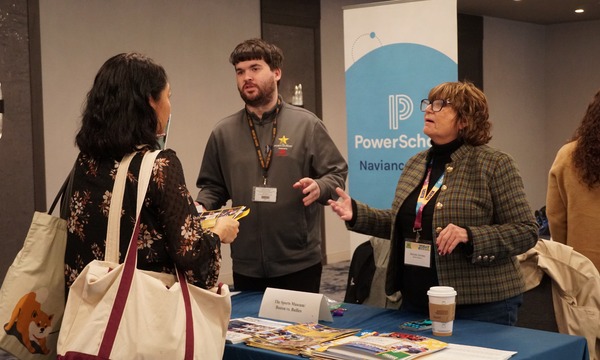 Conference attendee speaking with exhibitors at a booth display while reviewing conference materials and handouts