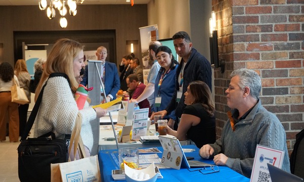 Conference attendee talking with exhibitors seated at display tables in a busy exhibitor hall.