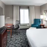 Modern hotel guest room with a king-size bed, flat-screen TV on a dresser, workspace with an ergonomic chair and desk lamp, blue upholstered chair, bedside lighting, and a large window with curtains.