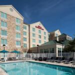 Exterior view of a Hilton Garden Inn hotel with a large outdoor swimming pool, lounge chairs, and patio umbrellas on a sunny day.