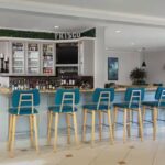 Bright hotel bar with blue bar stools, beverage displays, countertop seating, and a casual gathering space.