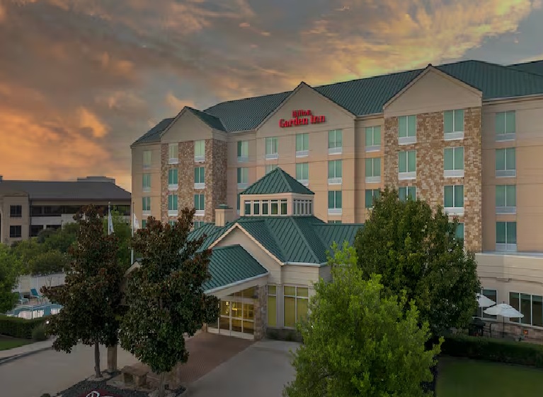 Front entrance of a Hilton Garden Inn hotel surrounded by trees and landscaping, photographed at sunset under a colorful sky.